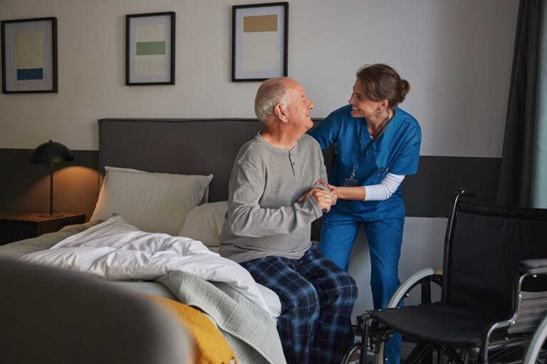 A middle-aged adult gently assisting an older woman as they walk together through a living room filled with natural light.